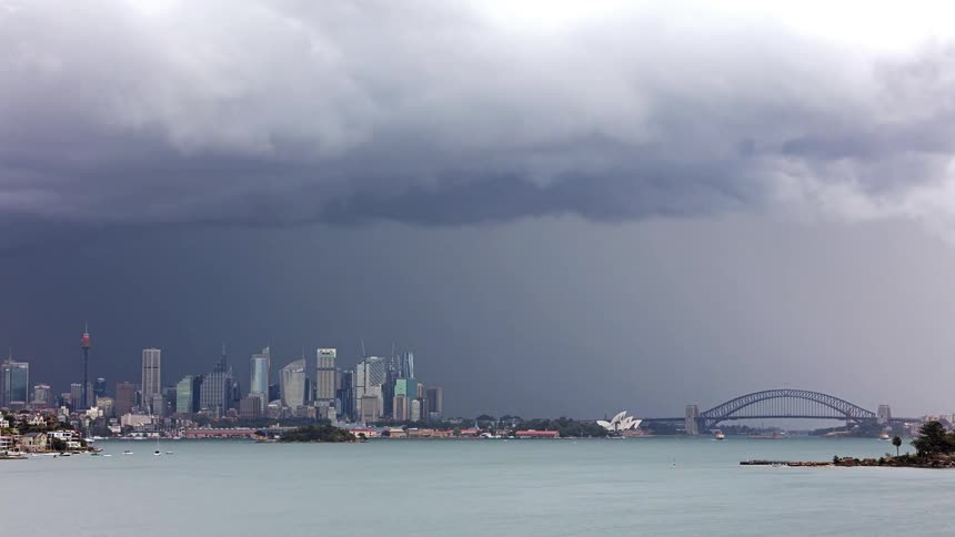 A view of a city skyline with many tall buildings and a large bridge over a body of water as thick grey clouds move across the sky
