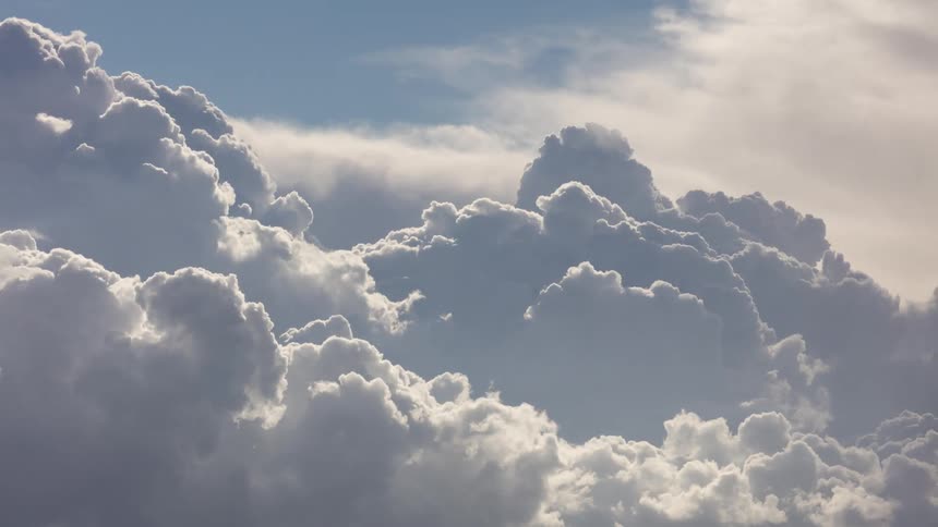 Large white cumulus clouds with soft textures and gray shadows fill the sky as sunlight glows through the gaps between the dense formations