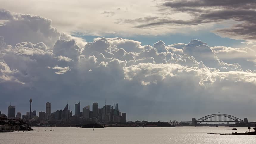 A view of a city skyline with many tall buildings and a large bridge over a body of water as thick white clouds move across the sky
