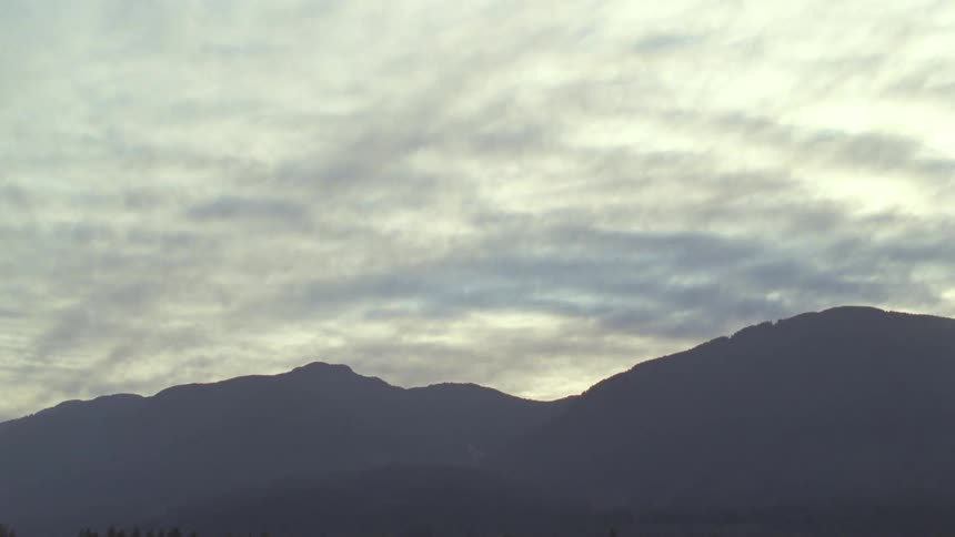 A dark jagged mountain range stretches across the lower third of the frame silhouetted against a pale skya dense layer of rippling overcast clouds moves slowly above the peaks filtering a soft and muted light over the landscape