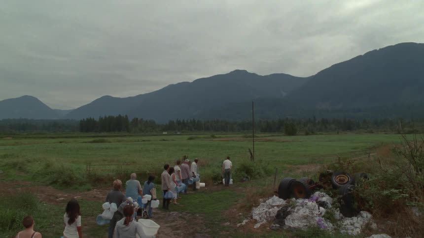 A group of people carrying buckets wait in a long line across a vast field with mountains in the background