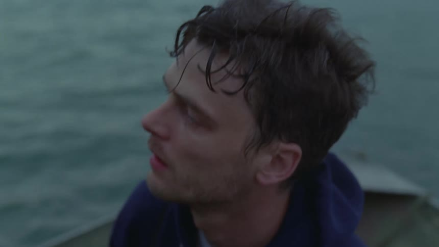 A close view of a man with wet hair wearing a blue hooded sweatshirt sitting on a boat and looking out over the water under a cloudy sky