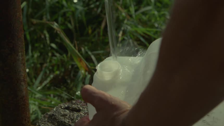 A person holds a large white plastic container under an outdoor spigot to fill it with waterthe water splashes against the opening of the jug and the surrounding grass as the container is tilted and moved