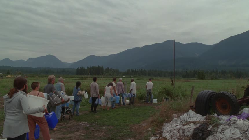 Under a heavy overcast sky a diverse group of people stands in a patient line across a grassy rural field to fill plastic jugs with water from a single ground source framed by a vast and somber mountain range