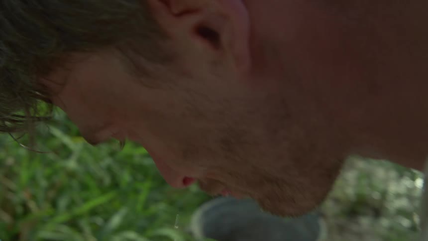 Young adult man wearing a white shirt washing his face on a faucet next to a road with trees and a mountain on the background outdoors during day time