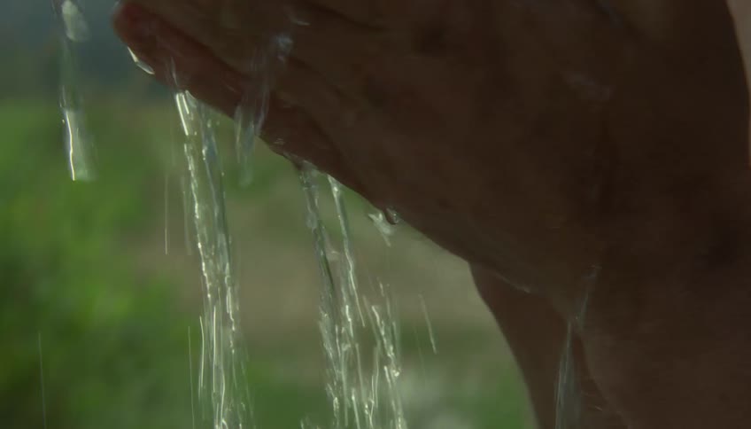 A man with a dirtstreaked face splashes water onto his eyes and forehead rubbing his skin as droplets glisten in the bright outdoor light