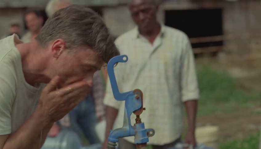 Man washing his face with a hand pump water gushing out in the open air while other people behind him hold gallon jugs