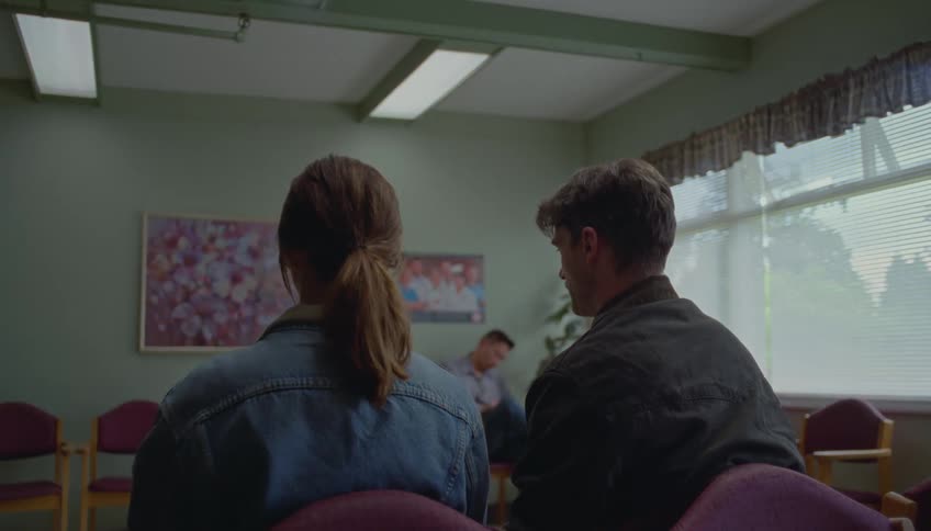 A man and woman sit silently in a somber waiting room their backs to the camera as another man sits alone in the background with his head bowed under the dim glow of overhead fluorescent lights