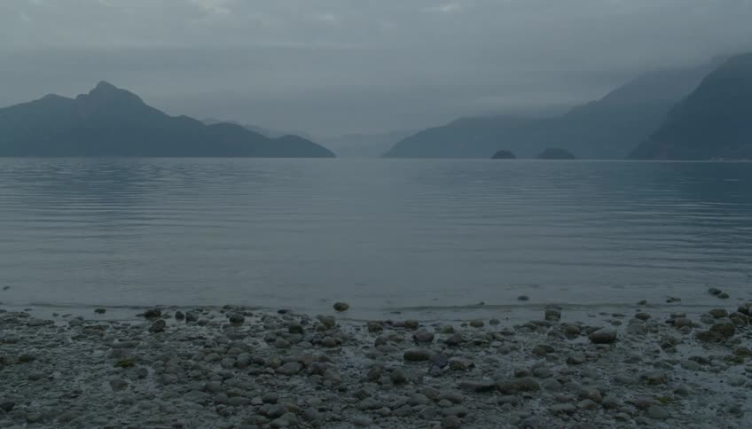 A calm lake at sunset with small rocks lining the shoreline a mountain range in the background and a soft fog settling over the scene
