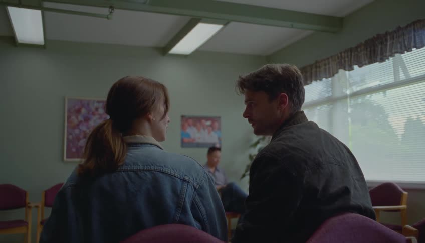 A young man and woman sit closely in a waiting room sharing a brief moment of conversation before she stands up and walks away the man watches her leave with a look of concern or anticipation as he remains seated in the quiet office