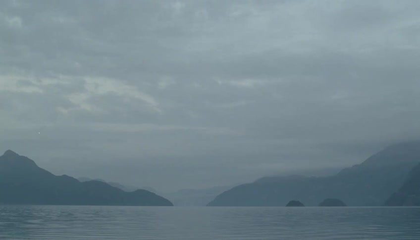 A wide shot captures a serene lake surrounded by dark mountain silhouettes under a heavy overcast sky