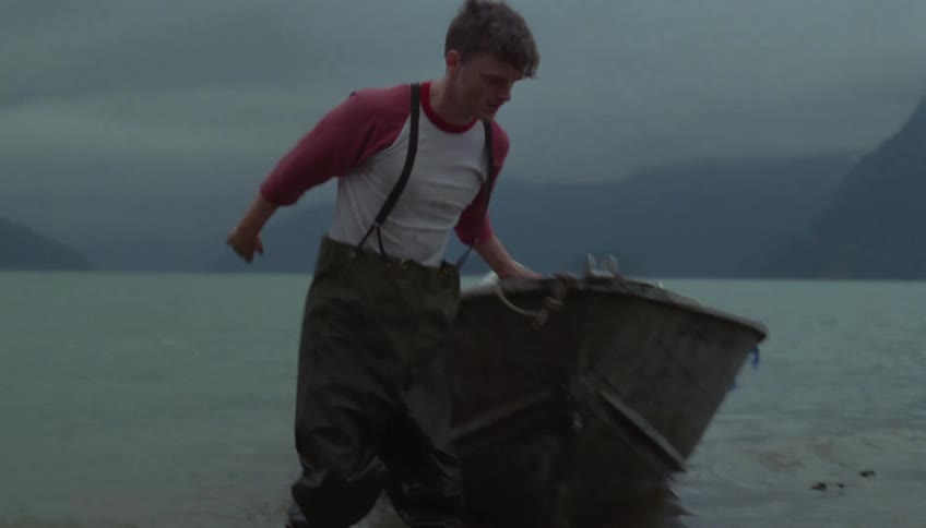 A man wearing chest waders and a redsleeved shirt pulls a small motorboat onto a rocky shorethe scene takes place under a heavy overcast sky with misty mountains rising from the water in the background