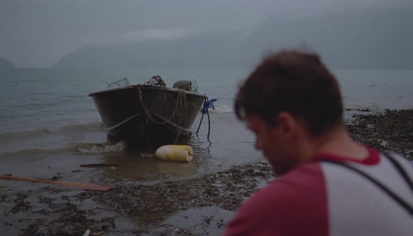 A man sits on a muddy rugged shore with a small boat anchored nearby on a foggy overcast day