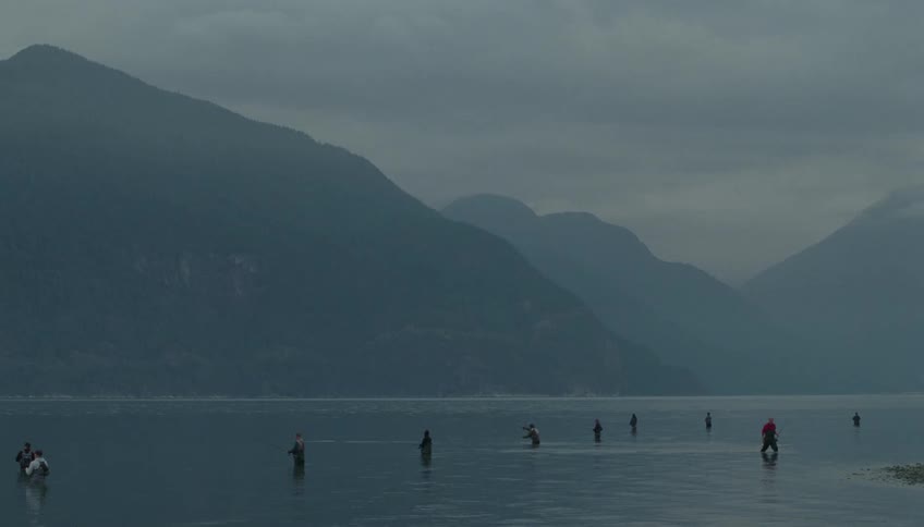 Several fishermen stand waistdeep in a calm body of water casting their lines against a backdrop of misty dark mountainsthe overall atmosphere is quiet and moody characterized by a heavy overcast sky and the still greyishblue tones of the landsca