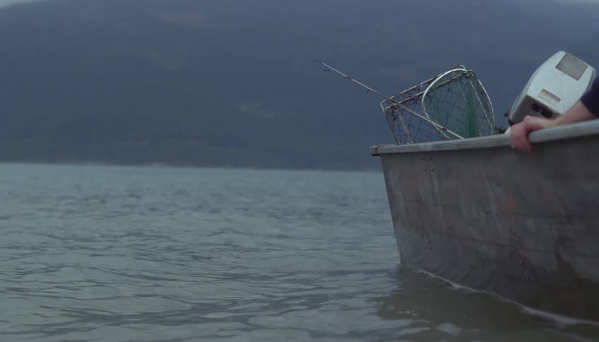 A view from the side of a small boat on a calm body of water with fishing gear and a motor visible against a background of misty mountains