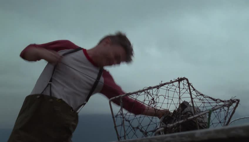 A young man angrily drags the engine of a fishing boat across the beach trying to get it started it fails again and he storms off across the sand under a cloudy gray sky