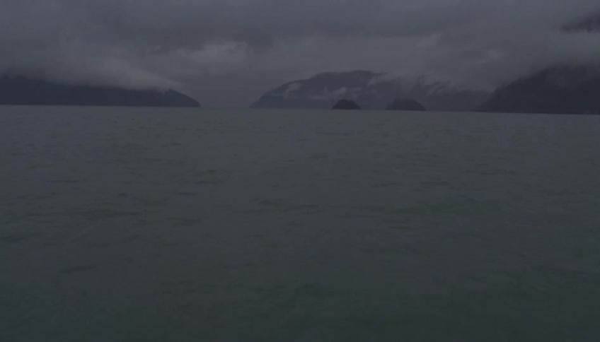 A view across a vast and dark body of water shows misty mountains and low gray clouds in a calm and somber atmosphere