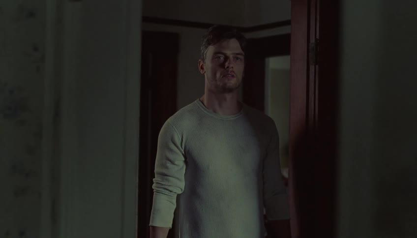 A man wearing a light colored long sleeve shirt stands in a dimly lit doorway and looks forward with a serious expression
