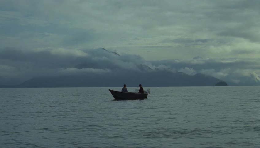 Small boat with two people moving across calm waters with cloudcovered mountains in the background