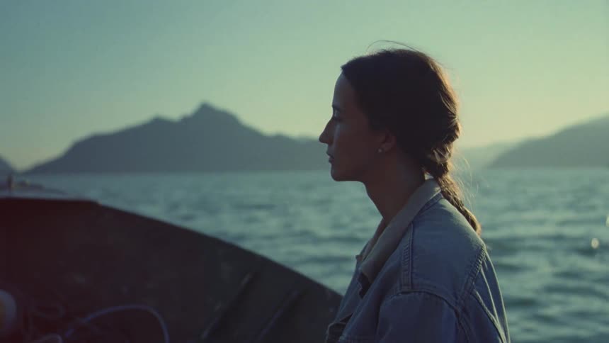 Woman sailing in a small boat across calm ocean waters toward distant mountains at sunset