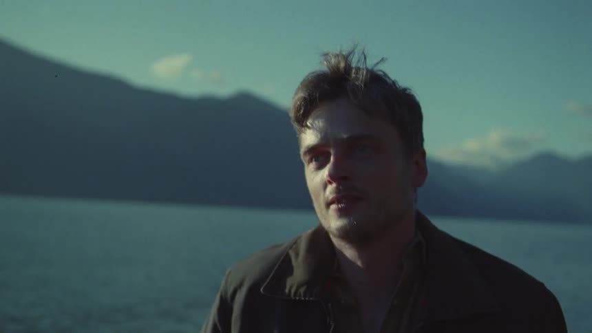 Man in the foreground facing a lake with mountains in the background under natural light
