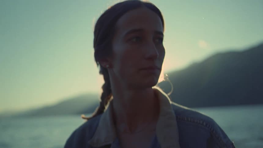 Standing on a moving boat a young woman with a braid gazes out at the shimmering water and distant mountain peaks as the warm glow of the setting sun creates a soft lens flare across the cinematic landscape