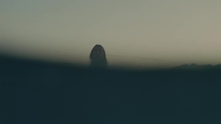 A young woman walks through the shallow ocean water near the coastline at sunset the warm light reflecting off the waves everything from her waist down is submerged as she moves slowly through the calm glowing water