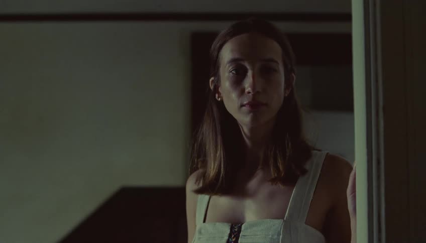 A young woman stands in a dimly lit doorway gazing forward before turning to walk away