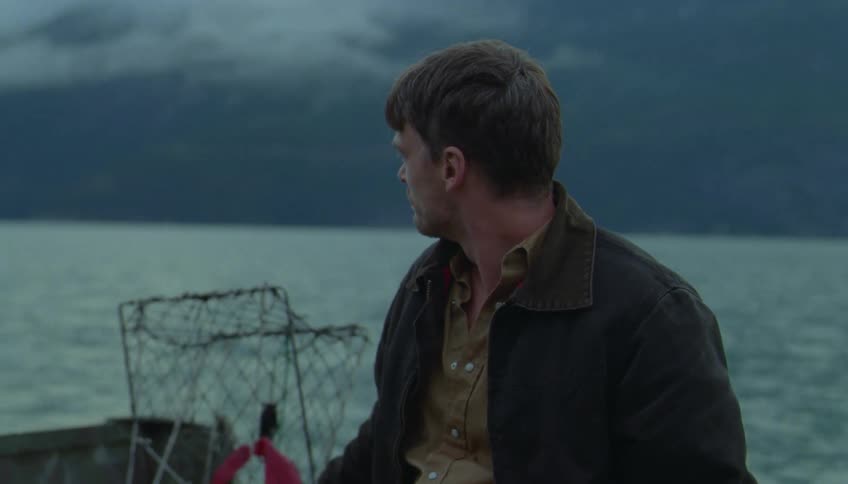 A man in a dark jacket stands on the deck of a fishing boat glancing out over the choppy gray water toward a misty mountainous coastline under a heavy overcast sky