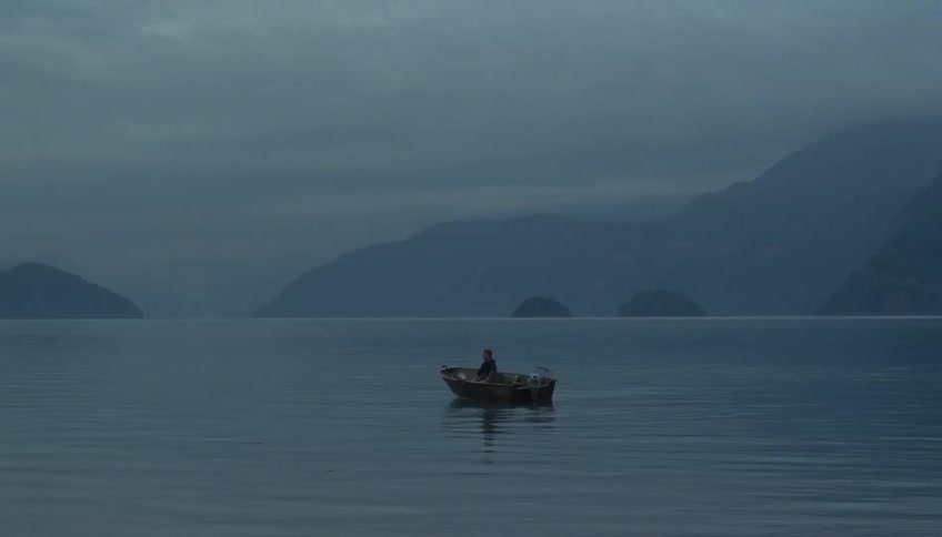 A man lying on a fishing canoe floating on a lake in a foggy atmosphere at sunset with a mountain range in the distance