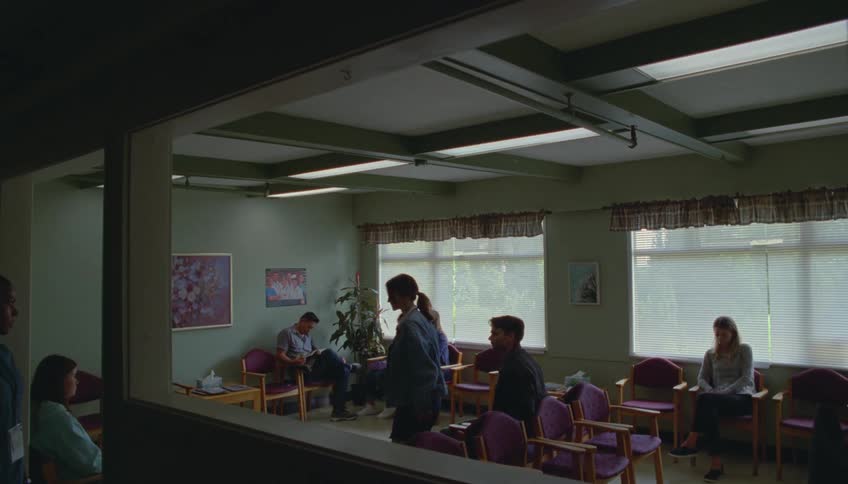 People wait seated in a waiting room with aligned chairs and large windows