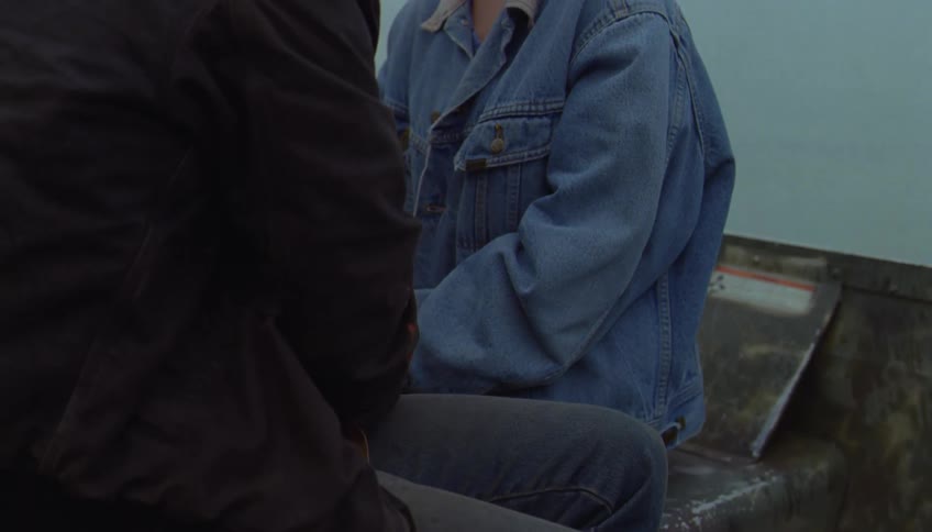 A man in a black jacket sits on a boat and speaks to a woman wearing a blue denim jacket while they look at each other with the calm grey water of the sea and a cloudy sky in the background