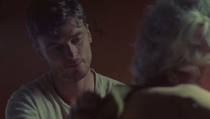 A man sits in a dimly lit room and looks up with a thoughtful expression while engaging in a quiet conversation with an older personhe then leans forward with a gentle smile to carefully wash or dab the older persons shoulder with a sponge
