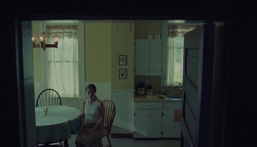 Viewed through a dark doorway a solitary woman sits at a kitchen table her expression shifting in the quiet stillness of the sunlit room