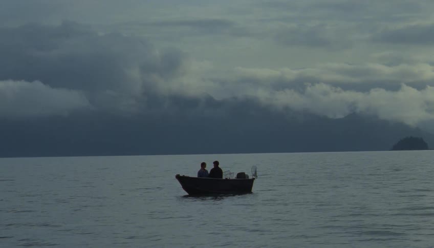 Two people float in a small boat on the water set against a backdrop of misty mountains and a dark cloudy sky