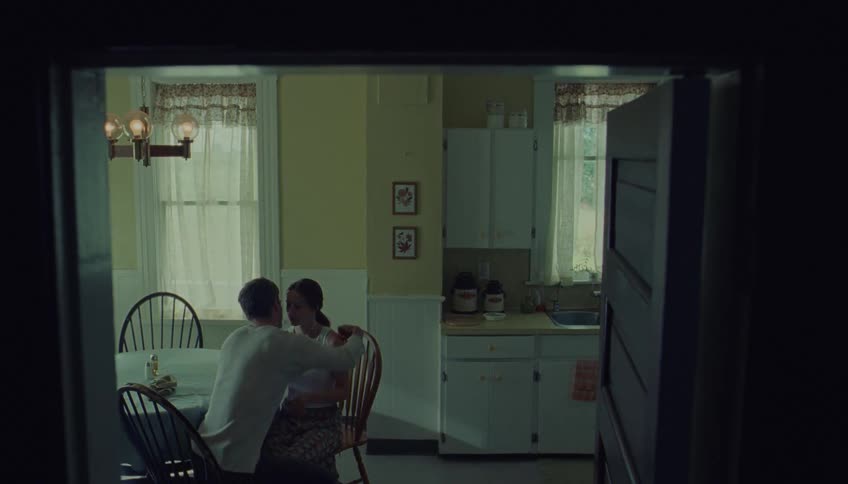 A couple sits at a kitchen table and shares a somber embrace viewed through a dark doorway