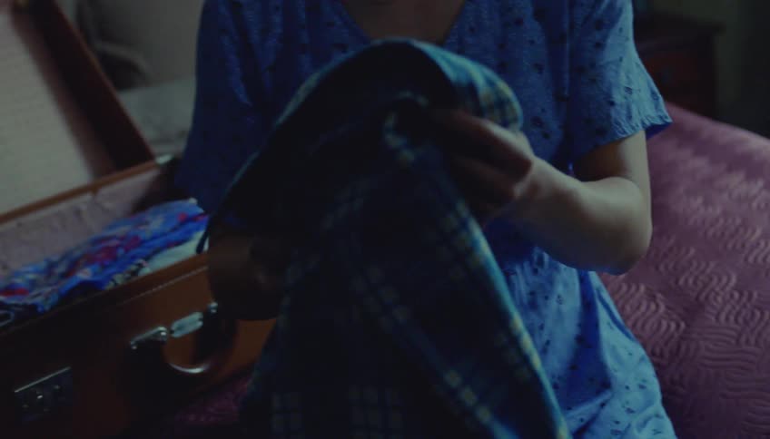A woman with dark hair in a braid wearing a blue floral shirt sits on a bed and carefully folds a blue patterned garment next to an open suitcase in a dimly lit room