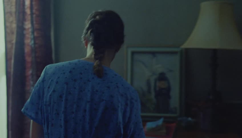 A woman with a dark braid wearing a blue patterned shirt stands in a room packing a suitcase on a bed