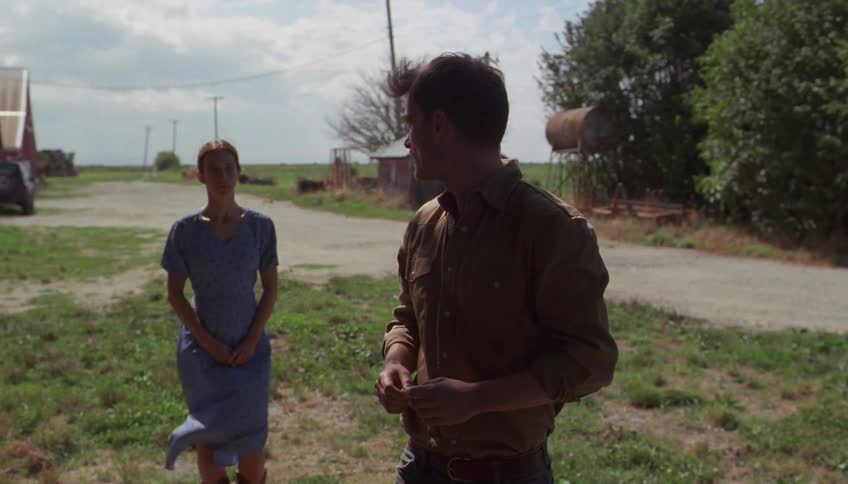 Man and woman talk on a rural road surrounded by fields and farm structures