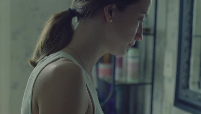 A woman turns on both the hot and cold faucets of a bathroom sink finding no running water before turning them off and looking with a somber expression