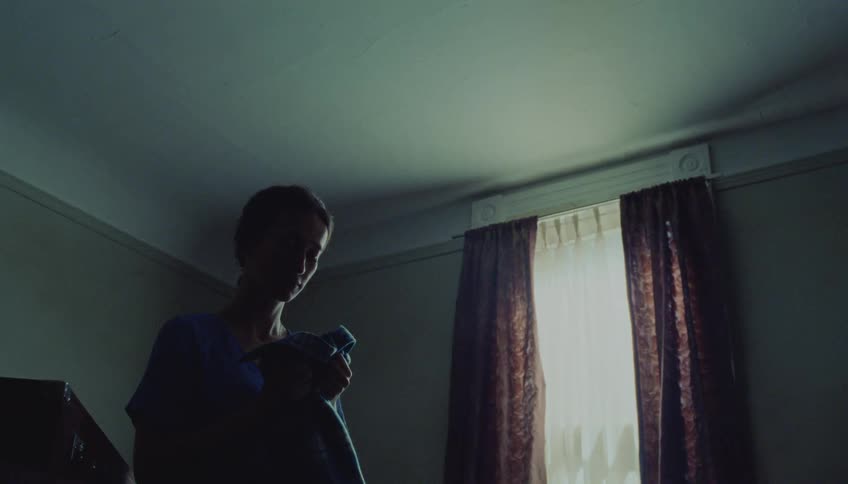 Young adult woman wearing a blue dress standing on a bedroom smelling a shirt with a sad facial expression indoors during day time