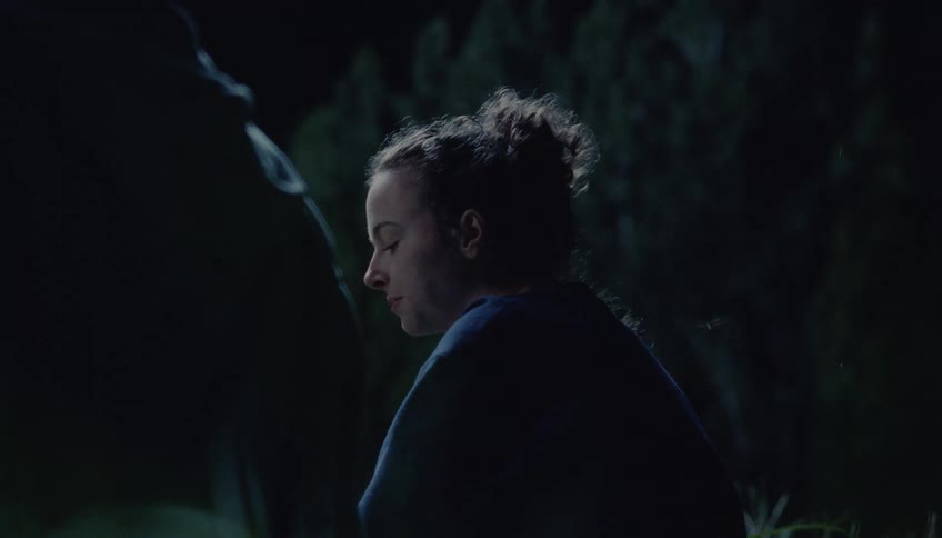 A young woman with her hair in a bun sits outside at night wearing a blue sweatshirt as she looks off into the distance