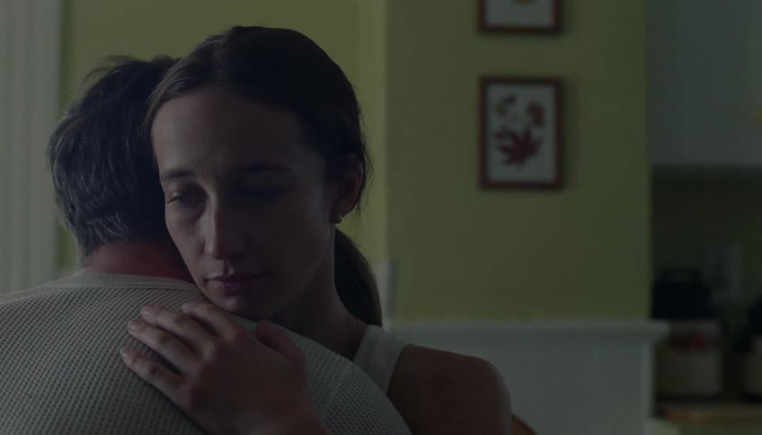 A young woman sits indoors looking sad as a young man leans in and hugs her gently