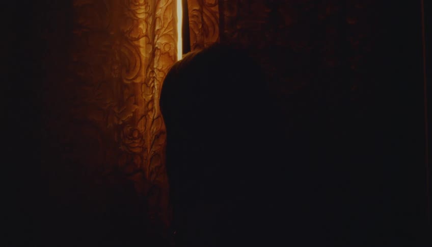 A person stands in a dark room and pulls back heavy patterned curtains to peek through a small gap where bright golden light shines through from the other side illuminating their silhouette and the texture of the fabric