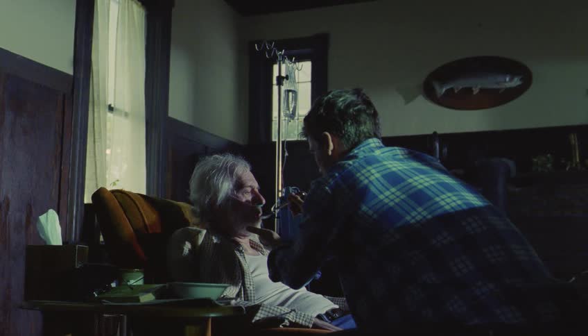 A man in a blue plaid shirt sits beside an elderly man in a chair and gently feeds him with a spoon in a dimly lit room with medical equipment and a fish mount on the wall