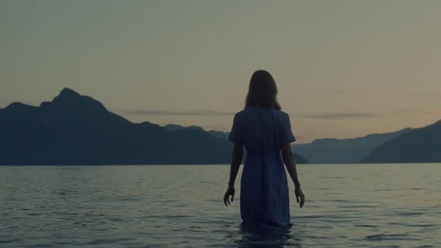 Woman seen from behind standing waistdeep in a lake with mountains in the background at dusk