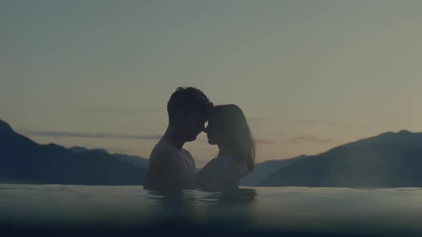 A couple embraces in a pool at twilight silhouetted against a backdrop of distant mountainsthe camera moves rhythmically above and below the waters surface creating a dreamy atmospheric perspective of their intimate moment