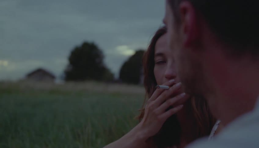 Woman holds a cigarette near her mouth while talking to another person in a rural area at dusk