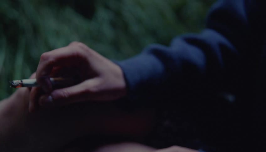 A close view shows the hand of a woman wearing a blue sleeve as she holds a burning cigarette while sitting outdoors near green foliage