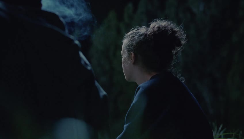 A woman smoking a cigarette is sitting next to another person in the woods
